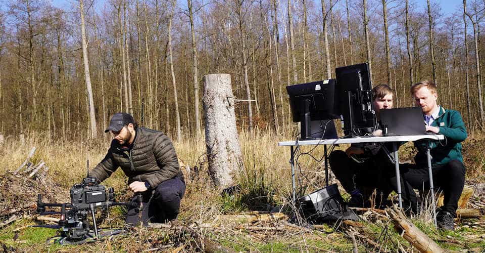 Ein Mann sitzt vor einer Drohne, zwei Männer sitzen an einem Tisch mit mehreren Monitoren