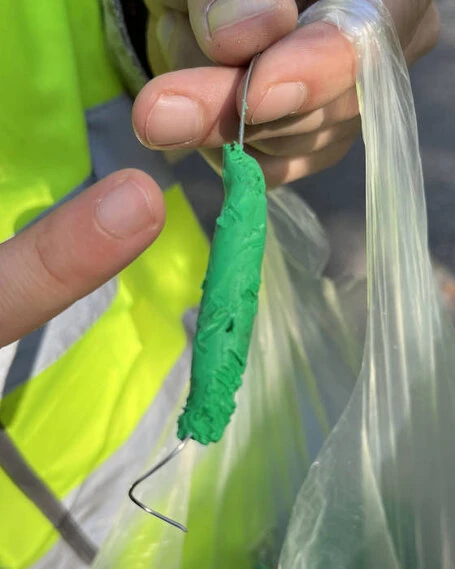 Hand hält eine grüne Raupe aus Knetmasse an einem Draht, im Hintergrund eine durchsichtige Plastiktüte und reflektierende Kleidung.